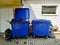 Two blue dumpsters. Made of plastic, with lids. Lying next to the garbage, the waste. Stand near the building on the tile