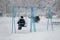 Two boys ride on a blue swing in a winter park during a snowfall. Back view