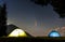 Two brightly lit tourist tents on forest clearing in mountains with starry sky and C/2020 F3 NEOWISE comet with light tail