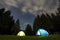 Two brightly lit tourist tents on green grassy forest clearing among tall pine trees on clear dark blue starry sky background.