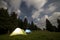 Two brightly lit tourist tents on green grassy forest clearing among tall pine trees on clear dark blue starry sky background.