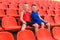 Two brothers of the child sit on red seats on the sports stand of the stadium