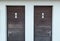 Two brown wooden toilets doors with man and woman symbol on the door, a white plastered wall surrounds the doors, a green dial