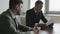 Two businessmen in suits are sitting at a table in front of the window and learning a new contract.