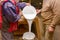 Two carpentry workers pour wood glue from a large plastic bucket into another empty