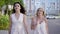 Two charming girls in white stylish dresses talking lively on background of street in summer.