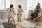 Two charming little girls in their pajamas are having fun jumping on a bed in a sunlit cozy bedroom with New Year`s tree