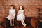 Two charming sisters dressed in beautiful dresses are sitting on the brown armchair on the background of a brick wall