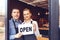 Two cheerful small business owners at entrance of newly opened pub restaurant holding open sign board