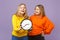 Two cheerful young blonde twins sisters girls in vivid colorful clothes hold round clock isolated on pastel violet blue