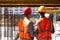 Two civil engineers dressed in orange work vests and helmets discuss the construction process on the building site near