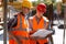 Two civil engineers dressed in orange work vests and helmets explore construction documentation on the building site