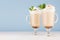 Two coffee drinks with whipped cream, green mint, cocoa powder for breakfast in glass with handle in pastel blue interior.