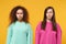 Two confused offended women friends european and african american girls in pink green clothes posing isolated on yellow