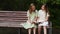 Two cute young girls in fashionable summer dresses reading interesting book sitting on old wooden bench in green park on
