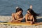 Two dark-haired girls in swimsuits and sunglasses lay on the yellow surfboard on the sandy beach near the sea on a sunny