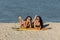 Two dark-haired girls in swimsuits and sunglasses lay on the yellow surfboard on the sandy beach near the sea on a sunny