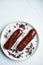 Two delicious eclairs with custard and glossy chocolate icing on a white ceramic plate on a marble background. Appetizing dessert