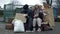Two dirty and poorly dressed homeless people, a man and a woman, sit by a pile of rubbish and eat and drink something. A
