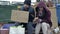 Two dirty and poorly dressed homeless people, a man and a woman, sit by a pile of rubbish with a handwritten SELL