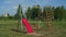 Two empty red rope swings swaying on metal playground in green meadow in warm summer day. Trees and house in background