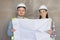 two engineers, a man and a woman in white helmets and protective vests, are standing in the room and holding a plan of