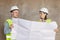two engineers, a man and a woman in white helmets and protective vests, are standing in the room and holding a plan of