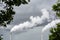 Two factory smokestacks emitting white smoke against a stormy sky with foliage in the foreground