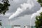 Two factory smokestacks emitting white smoke against a stormy sky with foliage in the foreground