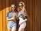 Two fashion beautiful girls in the marine clothing, shorts, stylish sunglasses standing near a wall of wooden planks. Drink a cool