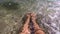 Two female feet are in clean transparent water on sandy beach on seashore