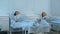 Two female patients on drips lying on beds in hospital ward being checked by two doctors