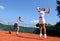 Two female tennis players playing doubles in the sun. One is leaping and stretching for the ball.