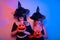 Two female witches hold pumpkins in their hands, covering their faces with a hat. Women in neon lights are celebrating a Halloween