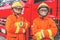 Two firefighters in protective clothing, helmets and mask against fire engine posing against fire truck background