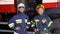 Two firefighters standing together, wearing uniform and protective helmets. Fire trucks in the background