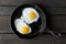 Two fried eggs in cast iron frying pan sprinkled with ground black pepper. Isolated on dark painted wood from above.