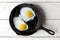 Two fried eggs in cast iron frying pan sprinkled with ground black pepper. Isolated on white painted wood from above.
