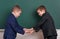Two friends handshake, elementary school boy near blank chalkboard background, dressed in classic black suit, group pupil, educati