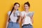 Two friends holding cash in hands, looking at camera with happy facial expression, ladies with dollars, female against yellow wall