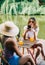 Two friends sitting on a pier lake, on summer day using mobile phone, communicate and drink lemonade