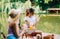 Two friends sitting on a pier lake, on summer day using mobile phone, communicate and drink lemonade