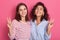 Two girlfriends having fun isolated over pink background, girls look at camera with toothy smile and showing victory sign,