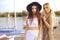 Two girls on the background of yachts, sailing boats are smiling, looking at the camera. Brunette in a hat and blonde in a dress