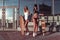 Two girls girlfriends posing summer city, background building glass windows, casual wear swimwear, hands longboard