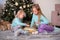 Two girls sisters draw fingers on pillow with sequins sit near Christmas tree.