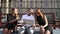 Two girls student and male student communicate on the bench in sunny weather using laptop