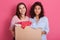 Two girls volunteering holding paper box with clothes for poor people, brunette ladies making donation, attractive females wearing