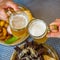 Two glass of beer in hand. Beer glasses clinking in bar or pub on table with food background