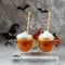 Two glass cups of pumpkin juice with painted faces on the background of Halloween pumpkins on a gray wall with ghosts